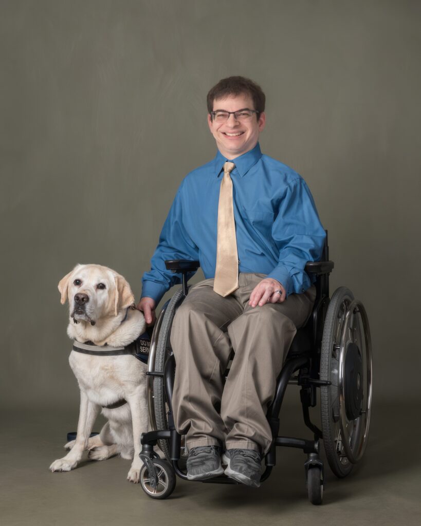 White person with short brown hair wearing a blue shirt, gold tie, and beige pants sits in a wheelchair. A service dog sits next to the wheelchair.