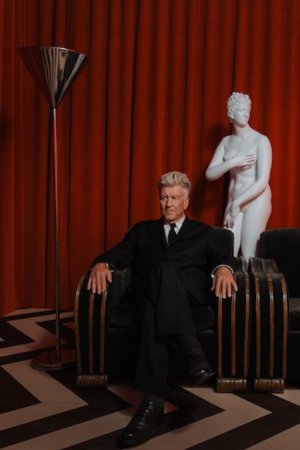 David Lynch sitting in the Red Room with red curtain background and black and white chevron floor.