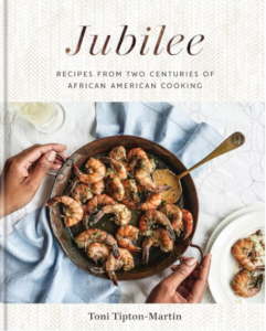 Front cover of Jubilee showing an overhead view of a serving dish of cooked shrimp in a sauce. A Black person's hands are on either side. A pale blue table linen is underneath.