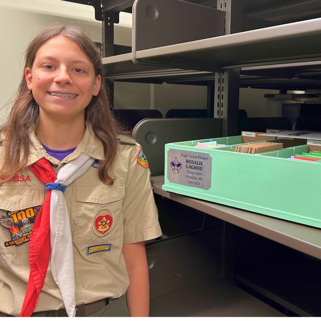 Photo of Eagle Scout Rosalie L. standing next to the seed library container she made and bearing a Scouting plaque with her name and troop on it.