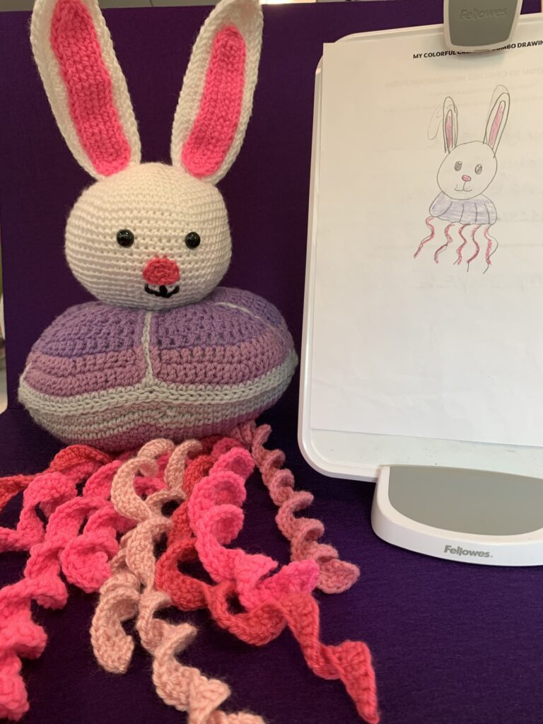 Crochet stuffy based on child's drawing which is also pictured. It's a purple jellyfish with pink tentacles and the head of a white bunny.