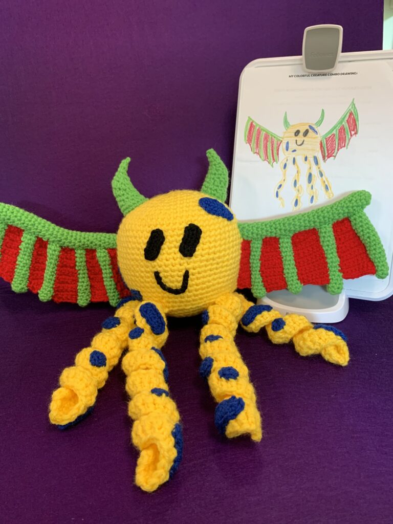 Crochet stuffy based on child's drawing which is also pictured. It's a yellow octopus with blue spots, and it also has green horns and red and green dragon wings.