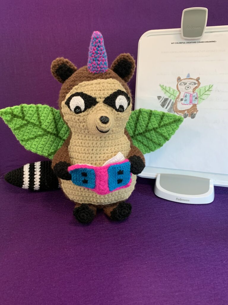 Crochet stuffy based on child's drawing which is also pictured. It's a racoon with leaf wings and a unicorn horn holding an open book.