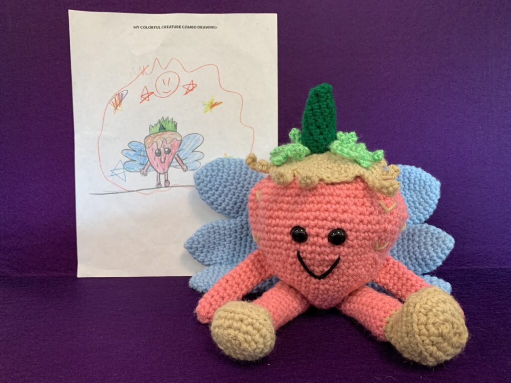 Crochet stuffy based on child's drawing which is also pictured. It's a pink strawberry with blue fairy wings and a green unicorn horn.