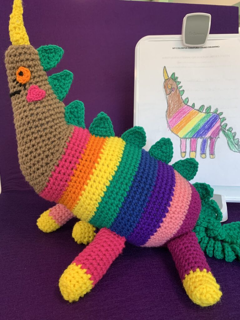 Crochet stuffy based on child's drawing which is also pictured. It's a rainbow unicorn with a brown neck and head and also has green dragon spines.