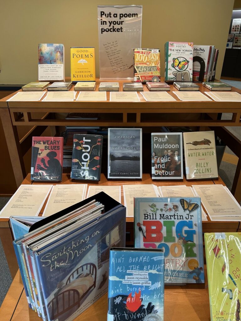 Display table featuring assorted poetry book covers and a central sign encouraging visitors to take poems from the selection.