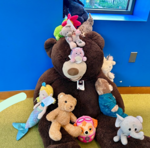 Ted, the large stuffed bear from Youth Services, is surrounded by various stuffies dropped off by young patrons.