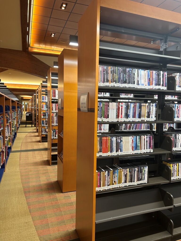 Tall wooden bookshelves filled with a variety of books line the carpeted floor of a well-lit library interior, with overhead lights brightening the aisles.